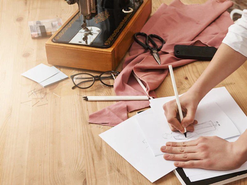 میز کار یک طراح حرفه‌ای لباس که با استفاده از مداد طرح‌های زیبایی روی کاغذ می‌آفریند و با سوزن و پارچه اثری هنری خلق می‌کند.